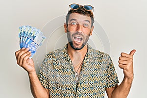 Handsome man with beard holding south african 100 rand banknotes pointing thumb up to the side smiling happy with open mouth