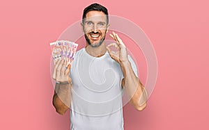 Handsome man with beard holding south african 50 rand banknotes doing ok sign with fingers, smiling friendly gesturing excellent