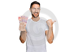 Handsome man with beard holding south african 50 rand banknotes pointing thumb up to the side smiling happy with open mouth
