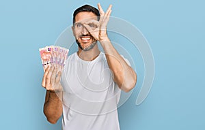 Handsome man with beard holding south african 50 rand banknotes smiling happy doing ok sign with hand on eye looking through
