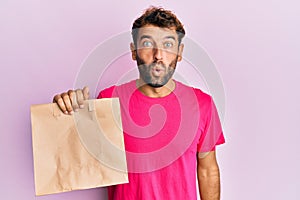 Handsome man with beard holding take away paper bag scared and amazed with open mouth for surprise, disbelief face