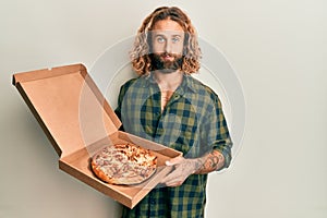 Handsome man with beard and long hair holding italian pizza relaxed with serious expression on face