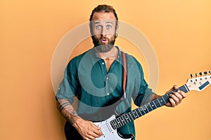 Handsome man with beard and long hair playing electric guitar in shock face, looking skeptical and sarcastic, surprised with open