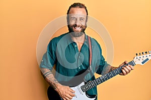 Handsome man with beard and long hair playing electric guitar smiling and laughing hard out loud because funny crazy joke