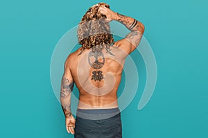 Handsome man with beard and long hair standing shirtless showing tattoos backwards thinking about doubt with hand on head