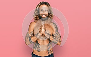 Handsome man with beard and long hair standing shirtless showing tattoos praying with hands together asking for forgiveness