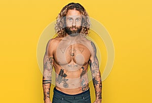 Handsome man with beard and long hair standing shirtless showing tattoos relaxed with serious expression on face