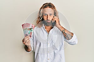 Handsome man with beard and long hair talking on the phone holding 100 new zealand dollars depressed and worry for distress,