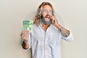 Handsome man with beard and long hair talking on the phone holding 50 shekels angry and mad screaming frustrated and furious,