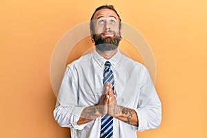 Handsome man with beard and long hair wearing business clothes begging and praying with hands together with hope expression on