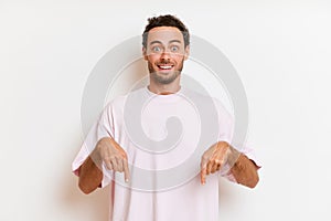 Handsome man with beard pointing down, smiles surprisedly wears white casual t-shirt stands isolated next to white