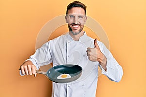 Handsome man with beard professional chef cooking fried egg smiling happy and positive, thumb up doing excellent and approval sign