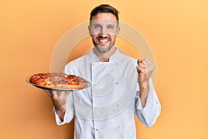 Handsome man with beard professional cook holding italian pizza screaming proud, celebrating victory and success very excited with