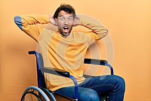 Handsome man with beard sitting on wheelchair crazy and scared with hands on head, afraid and surprised of shock with open mouth