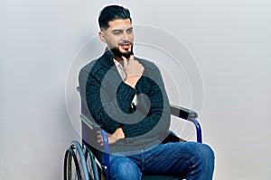 Handsome man with beard sitting on wheelchair with hand on chin thinking about question, pensive expression