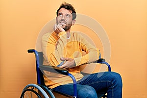 Handsome man with beard sitting on wheelchair with hand on chin thinking about question, pensive expression