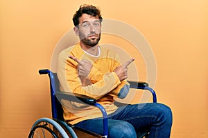 Handsome man with beard sitting on wheelchair pointing to both sides with fingers, different direction disagree