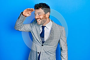 Handsome man with beard wearing business suit and tie very happy and smiling looking far away with hand over head