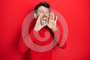 Handsome man with beard wearing casual red sweater shouting angry out loud with hands over mouth