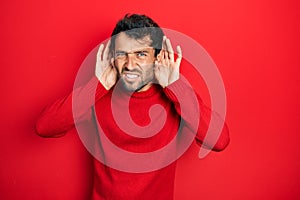 Handsome man with beard wearing casual red sweater trying to hear both hands on ear gesture, curious for gossip