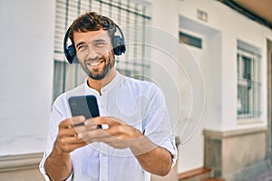 Handsome man with beard wearing casual white shirt on a sunny day smiling happy outdoors using smartphone listening to music