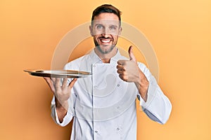 Handsome man with beard wearing chef uniform holding silver tray smiling happy and positive, thumb up doing excellent and approval