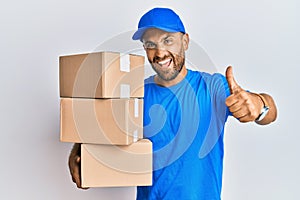 Handsome man with beard wearing courier uniform holding delivery packages approving doing positive gesture with hand, thumbs up
