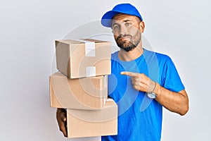 Handsome man with beard wearing courier uniform holding delivery packages pointing with hand finger to the side showing