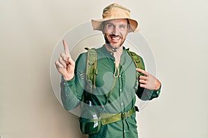 Handsome man with beard wearing explorer hat and backpack with a big smile on face, pointing with hand finger to the side looking