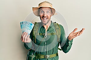 Handsome man with beard wearing explorer hat holding 100 brazilian reals celebrating achievement with happy smile and winner