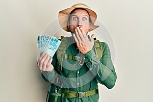Handsome man with beard wearing explorer hat holding 100 brazilian reals covering mouth with hand, shocked and afraid for mistake