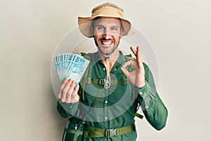 Handsome man with beard wearing explorer hat holding 100 brazilian reals doing ok sign with fingers, smiling friendly gesturing