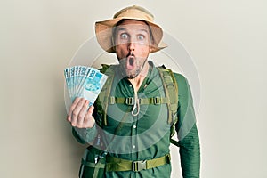 Handsome man with beard wearing explorer hat holding 100 brazilian reals scared and amazed with open mouth for surprise, disbelief