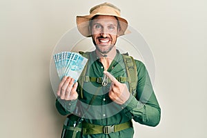 Handsome man with beard wearing explorer hat holding 100 brazilian reals smiling happy pointing with hand and finger