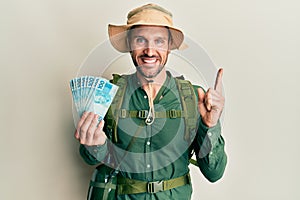 Handsome man with beard wearing explorer hat holding 100 brazilian reals smiling happy pointing with hand and finger to the side