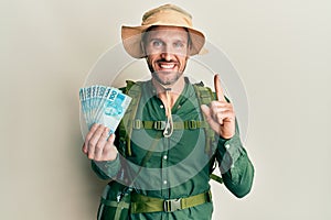 Handsome man with beard wearing explorer hat holding 100 brazilian reals smiling with an idea or question pointing finger with