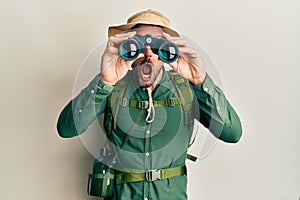 Handsome man with beard wearing explorer hat looking through binoculars afraid and shocked with surprise and amazed expression,