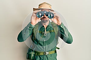 Handsome man with beard wearing explorer hat looking through binoculars puffing cheeks with funny face