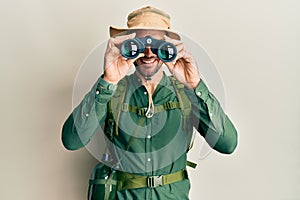 Handsome man with beard wearing explorer hat looking through binoculars smiling with a happy and cool smile on face