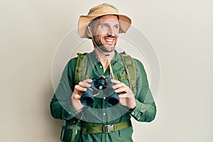 Handsome man with beard wearing explorer hat looking through binoculars smiling looking to the side and staring away thinking