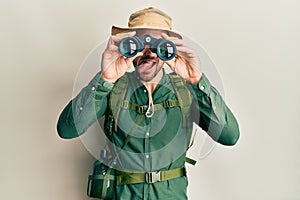 Handsome man with beard wearing explorer hat looking through binoculars sticking tongue out happy with funny expression