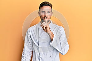 Handsome man with beard wearing professional cook uniform asking to be quiet with finger on lips