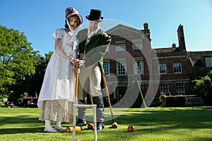 Handsome man and beautiful woman dressed in vintage clothing on lawn in front of stately home, playing croquet