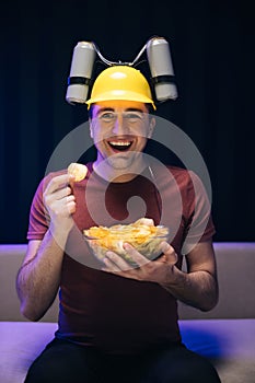 Handsome man with beer helmet on the head watching TV and eating potato chips on the couch at home. Young male sits on a
