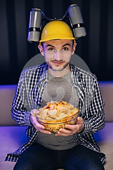 Handsome man with beer helmet on the head watching TV and eating potato chips on the couch at home. Young male sits on a