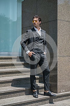 Handsome man in black formal suit and yellow sunglasses standing on the stairs and keeping his hands in his pockets.
