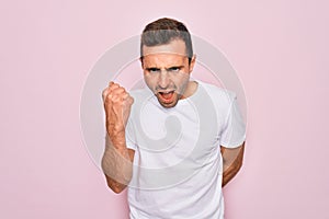 Handsome man with blue eyes wearing casual white t-shirt standing over pink background angry and mad raising fist frustrated and