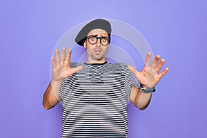 Handsome man with blue eyes wearing striped t-shirt and french beret over purple background afraid and terrified with fear