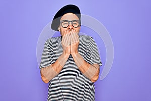 Handsome man with blue eyes wearing striped t-shirt and french beret over purple background laughing and embarrassed giggle