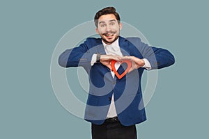 Handsome man in blue jacket and white shirt, standing and holding red heart shape and looking at camera with toothy smile.